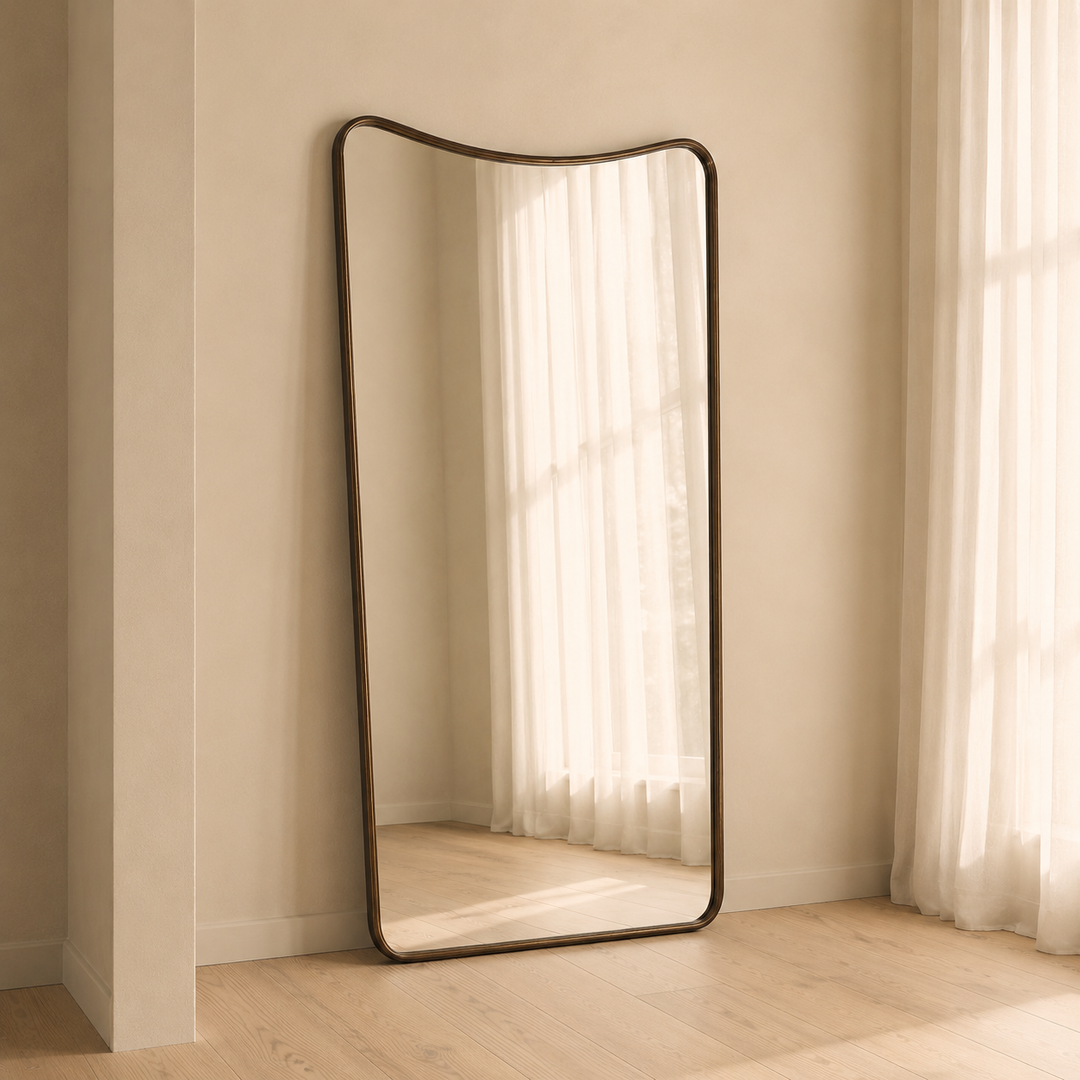 A tall floor mirror with a dark bronze curved frame leans against a beige wall on a light wood floor, reflecting sheer white curtains and natural light.