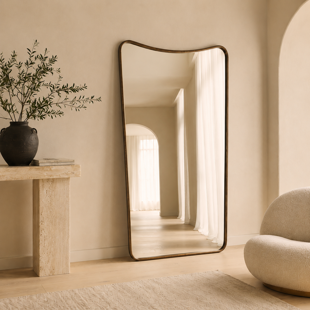 A tall, floor-standing mirror with an organic, wavy top border and a dark bronze frame, leaning against a beige wall in a minimalist, warm-toned room.