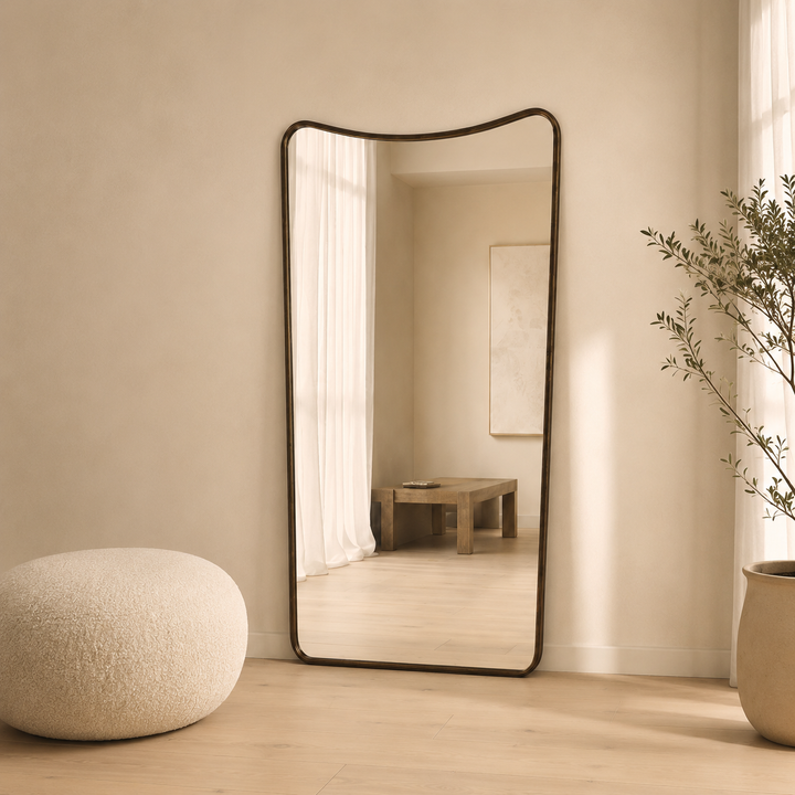 A tall floor mirror with a dark metal frame and curved top edge, leaning against a beige wall in a softly lit room. The mirror reflects a wooden table and curtains, while a round textured pouf sits on the floor to the left and a potted plant is on the right.