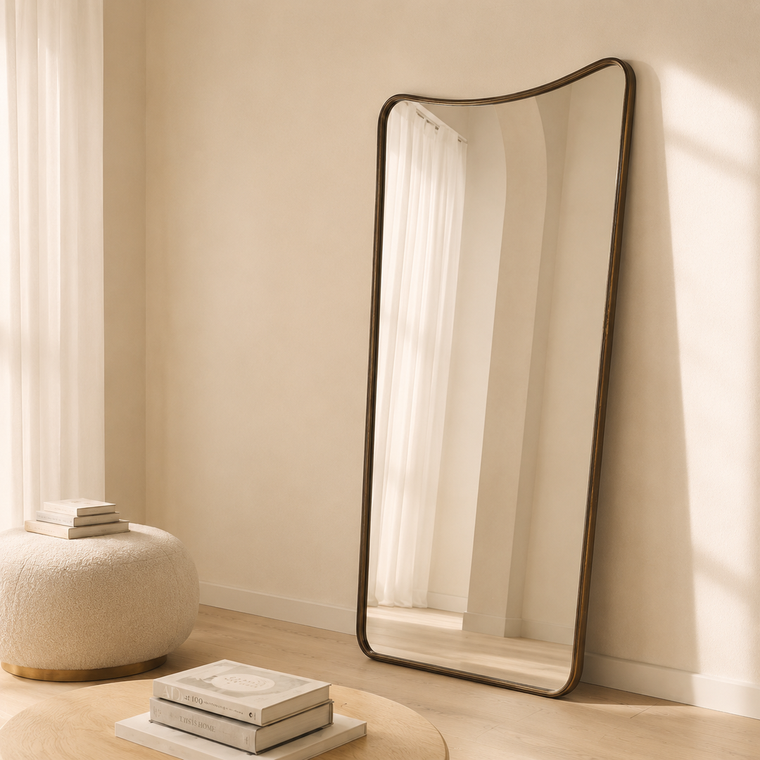 A tall, full-length floor mirror with a dark bronze curved frame leans against a beige wall in a sunlit room. The mirror reflects a white curtain, and the room features a textured cream pouf and stacked books on a light wood floor.