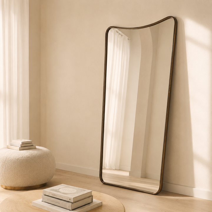A tall, full-length floor mirror with a dark bronze curved frame leans against a beige wall in a sunlit room. The mirror reflects a white curtain, and the room features a textured cream pouf and stacked books on a light wood floor.