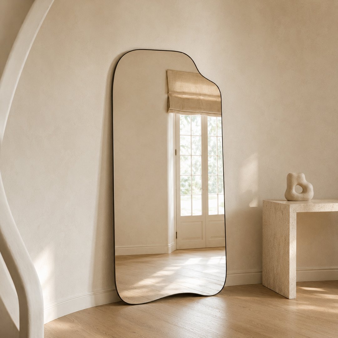 A full-length floor mirror with an organic, wavy top and bottom edge and a thin black frame, leaning against a textured beige wall in a minimalist room with light wood floors and a stone console table.