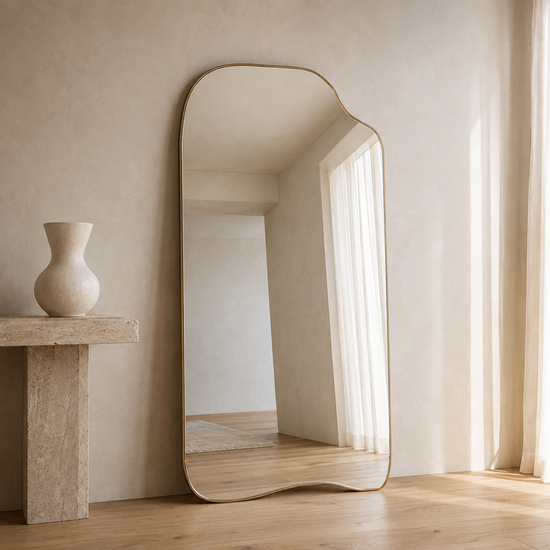 An asymmetrical full-length floor mirror with a wavy organic border and a thin gold frame, leaning against a textured beige plaster wall in a minimalist room with light wood floors and a stone side table.