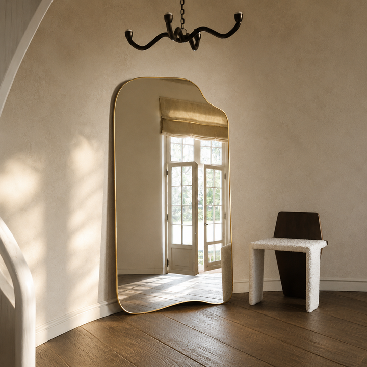 An asymmetrical full-length floor mirror with a gold frame leaning against a textured beige wall in a sunlit room with wooden floors and a small white bench.