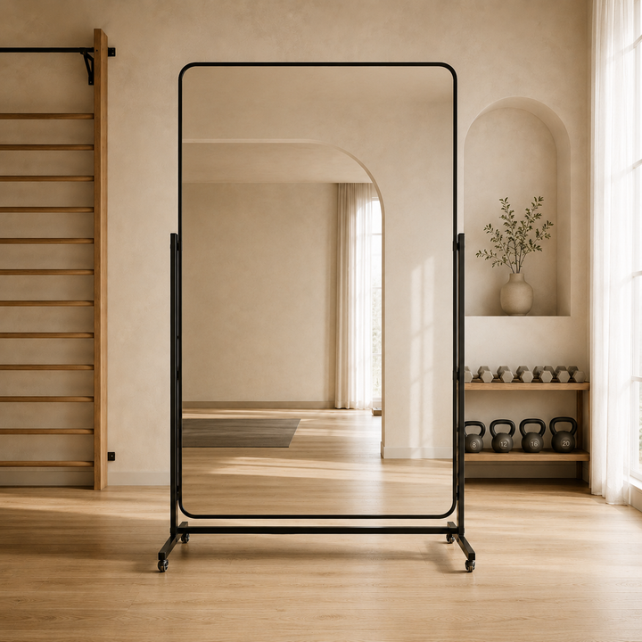 A tall freestanding floor mirror with a slim black metal frame and caster base, photographed in a warm, minimalist room with wood floors, an arched doorway, and shelving in the background.