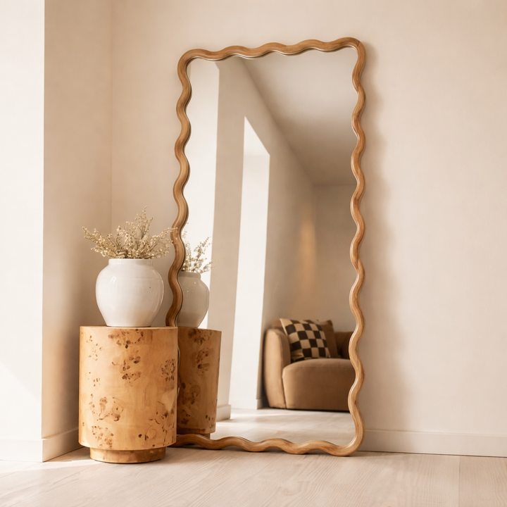Decorative mirror with wavy frame in a room setting with vases and a sofa.