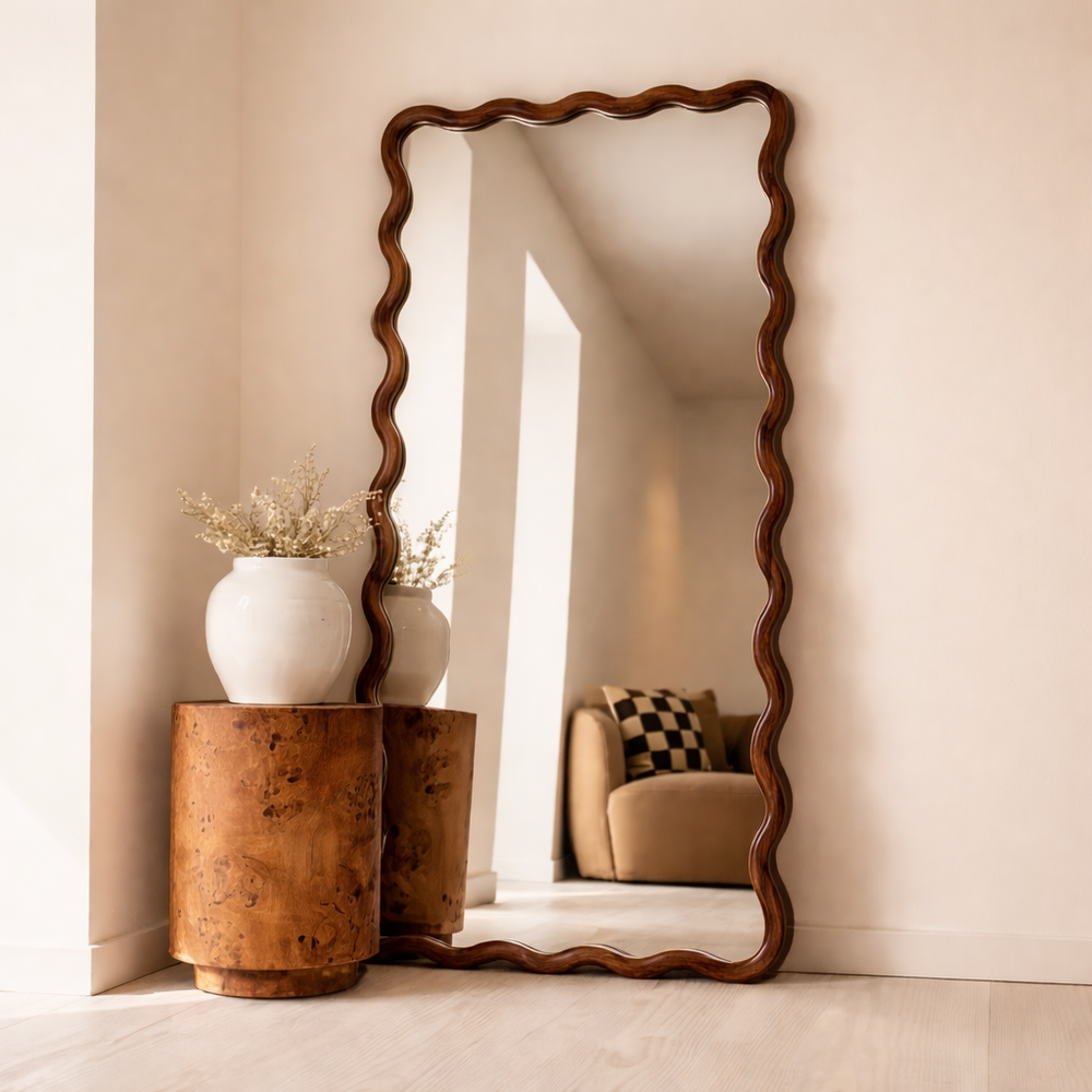 Wooden floor mirror with wavy design on a beige wall, with a side table and decorative items.
