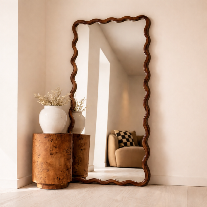 Wooden floor mirror with wavy design on a beige wall, with a side table and decorative items.
