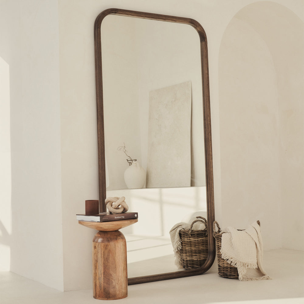 Large floor mirror with wooden frame leaning against a wall in a minimalistic room.