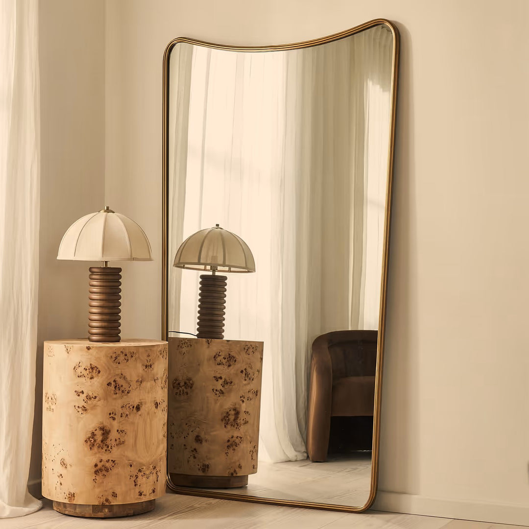 A tall, arched floor mirror with a brushed gold frame leans against a cream-colored wall in a styled interior. The mirror reflects a matching table lamp with a ribbed dark brown stem and a cream dome shade, set on a cylindrical burl wood side table.