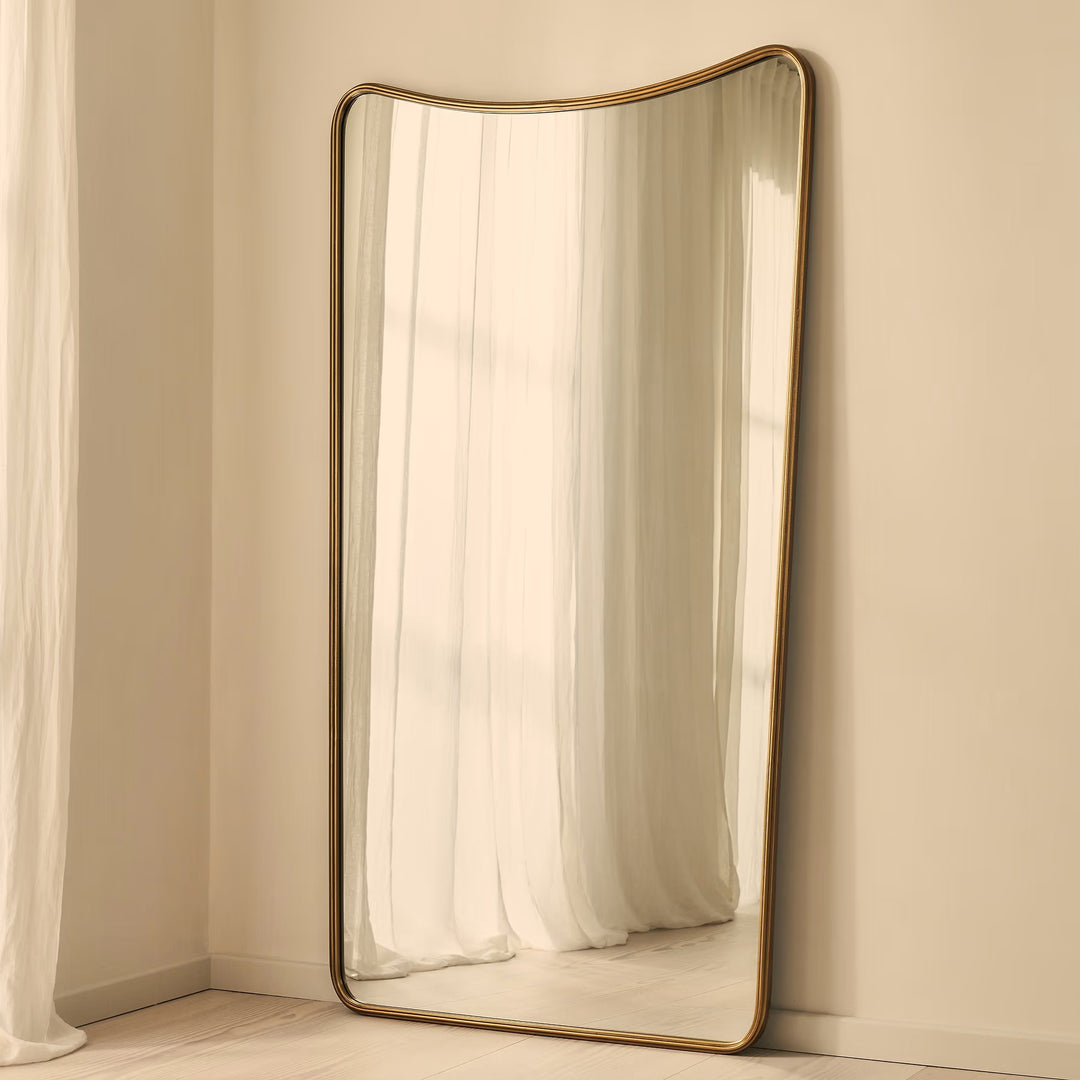 A full-length floor mirror with a curved top and a thin gold-toned metal frame, leaning against a beige wall in a softly lit room with white curtains.