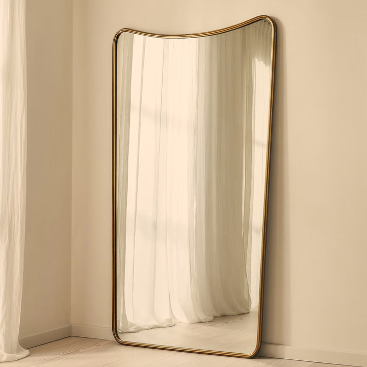 A full-length floor mirror with a curved top and a thin gold-toned metal frame, leaning against a beige wall in a softly lit room with white curtains.