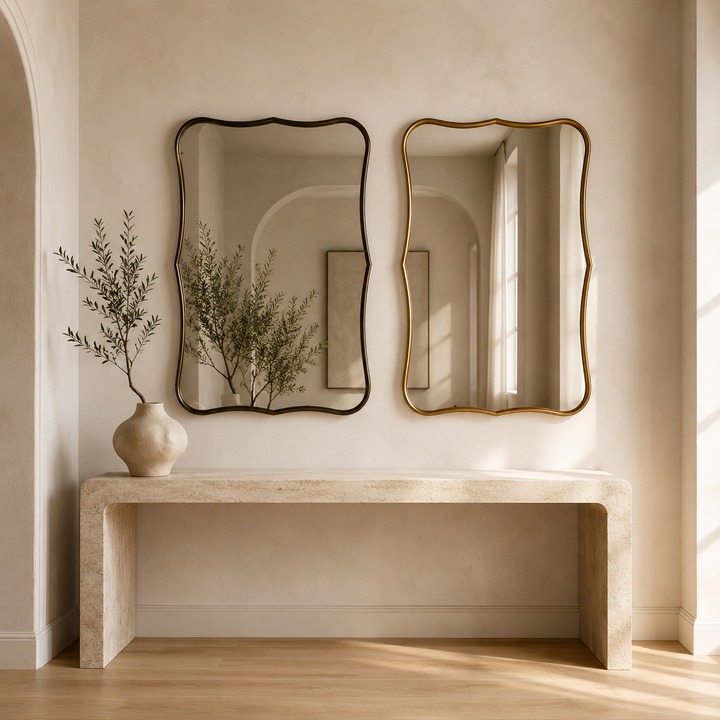 Two decorative wall mirrors with wavy scalloped edges hang above a stone console table in a warm beige interior. One mirror has a black frame and the other has a gold frame.