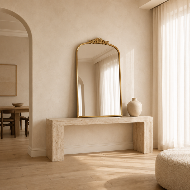 A tall rectangular wall mirror with an ornate gold frame and decorative crest at the top, leaning on a light stone console table in a warm, sunlit room. A cream ceramic vase sits to the right, with sheer curtains and a softly lit interior visible in the background.