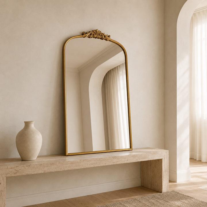 An arched-top gold-framed mirror with an ornate decorative crest at the top, leaning against a beige plaster wall on a light stone console table next to a ceramic vase.