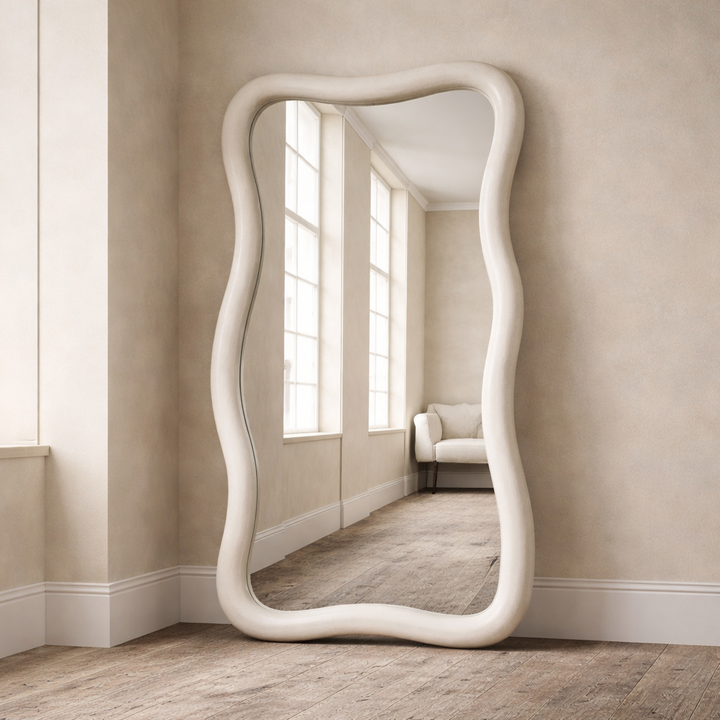 Decorative floor mirror with wavy frame in a room with large windows and a sofa.