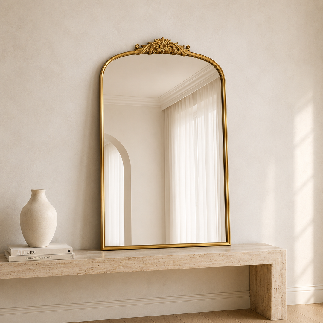 A tall arched mirror with an ornate gold frame and decorative crest at the top, leaning against a beige wall above a light stone console table. A small cream vase and books sit on the left side of the table.