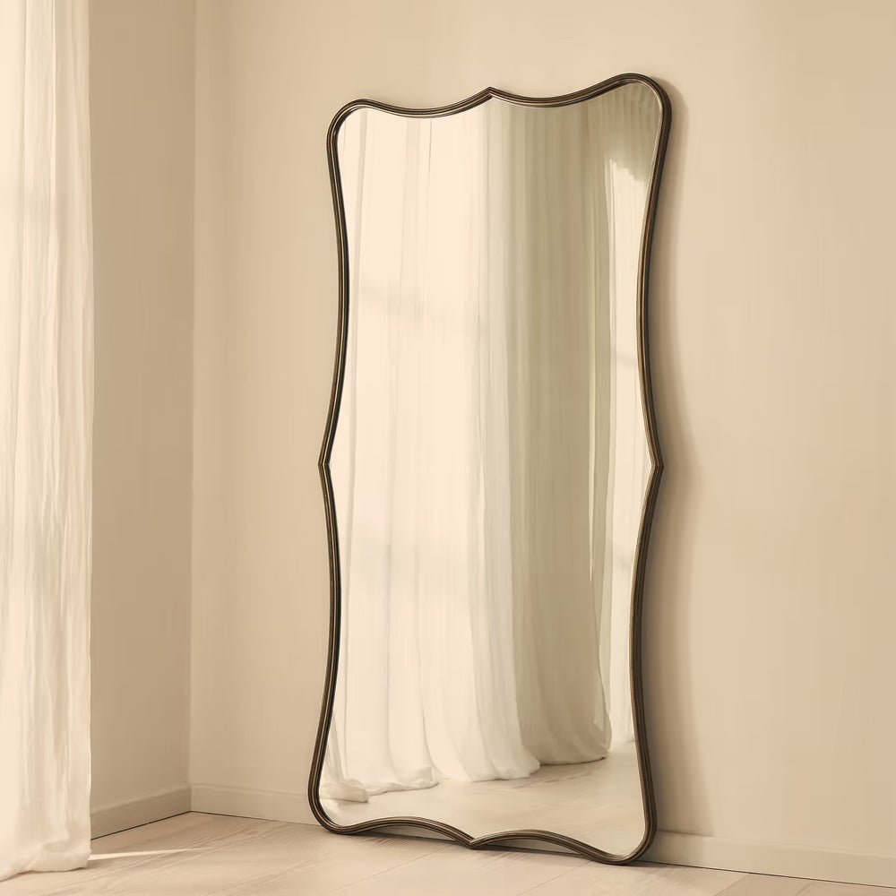 A full-length floor mirror with a dark, wavy scalloped frame leaning against a beige wall in a softly lit room.