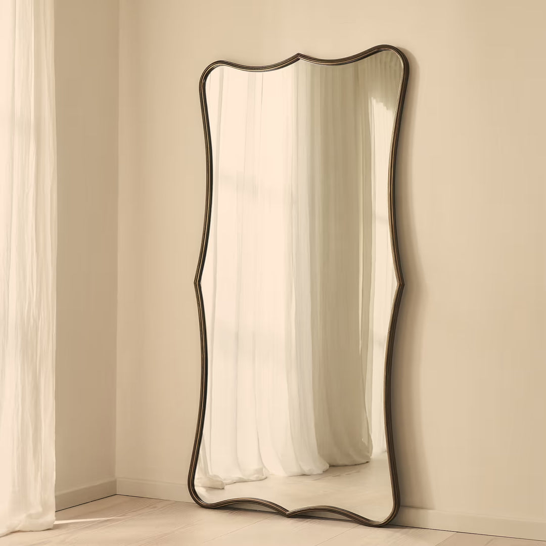 A full-length floor mirror with a dark, wavy scalloped frame leaning against a beige wall in a softly lit room.