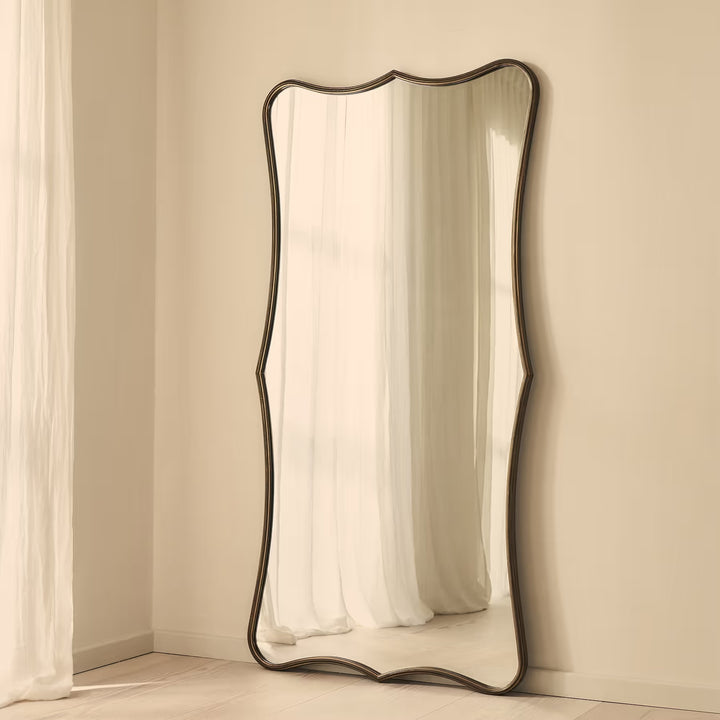 A full-length floor mirror with a dark, wavy scalloped frame leaning against a beige wall in a softly lit room.