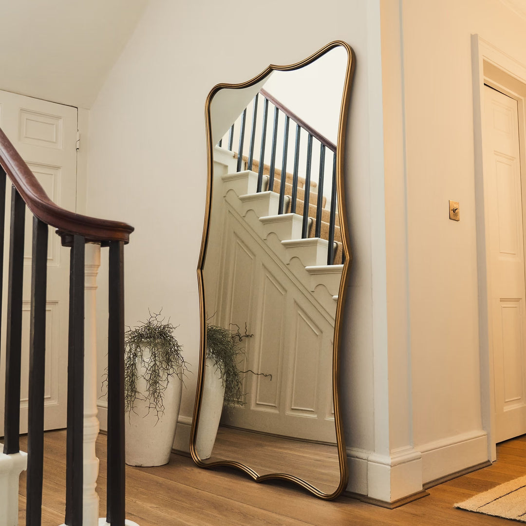 A full-length floor mirror with an ornate scalloped gold frame leans against a cream-colored wall in a staircase landing, reflecting a wooden staircase and white paneling.