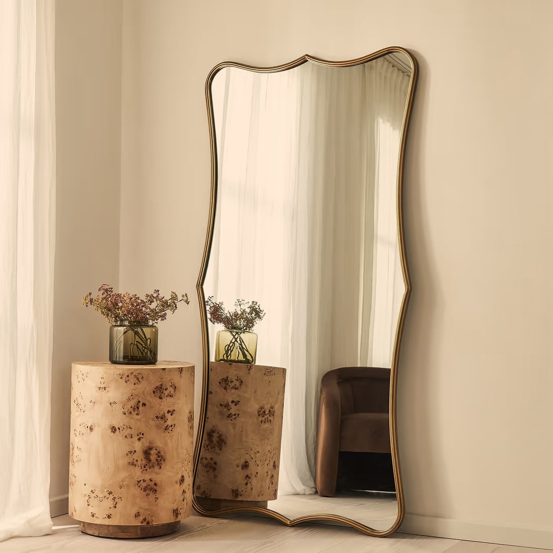A tall floor mirror with an organic wavy frame and a gold-toned metal border, leaning against a beige wall in a styled room next to a burl wood side table.