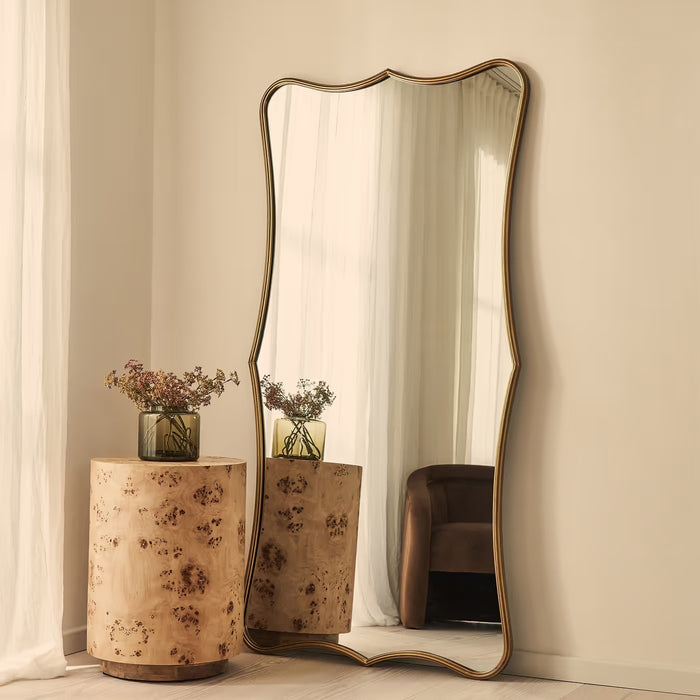 A tall floor mirror with an organic wavy frame and a gold-toned metal border, leaning against a beige wall in a styled room next to a burl wood side table.