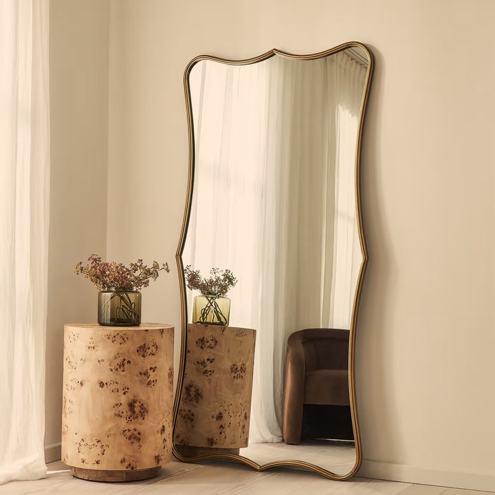 A tall floor mirror with an organic wavy frame and a gold-toned metal border, leaning against a beige wall in a styled room next to a burl wood side table.
