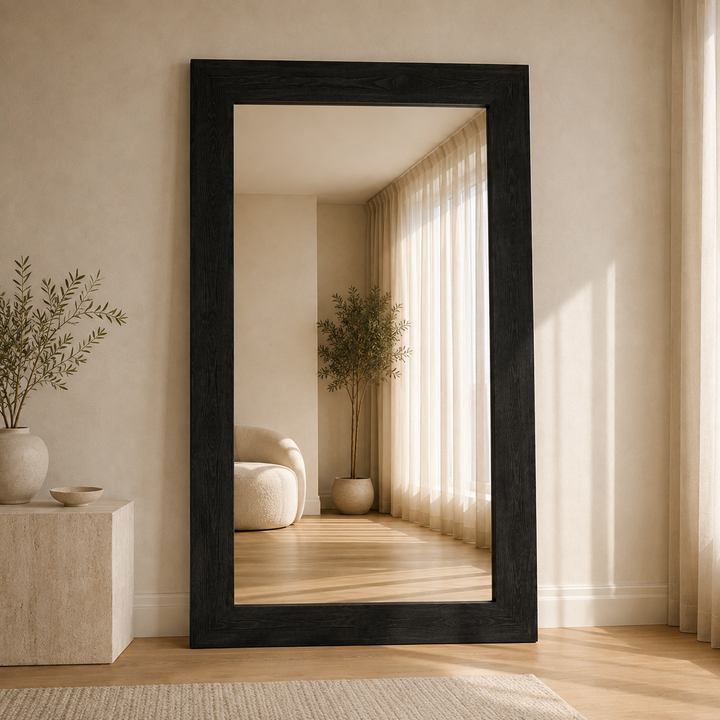 A tall, full-length floor mirror with a thick black frame, leaning against a beige wall in a minimalist, warm-toned room. The reflection shows a cozy interior with a plant, a cream-colored armchair, and sheer curtains.