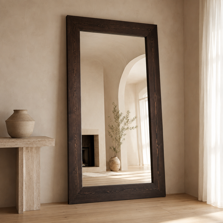 A tall rectangular floor mirror with a thick dark brown wooden frame, leaning against a textured beige plaster wall in a sunlit room. The mirror reflects an arched hallway with a vase of branches, and the room features a light wood floor and a stone side table with a ceramic vase.