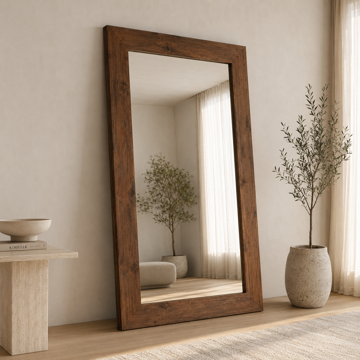 A tall floor mirror with a thick rustic dark brown wooden frame, leaning against a beige wall in a styled room with a potted tree and a side table.