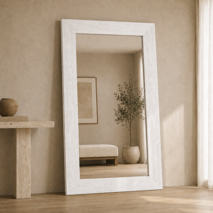 A tall floor mirror with a wide, distressed white frame stands leaning against a textured beige wall in a minimalist room. The mirror reflects a softly lit interior with a potted plant and a low bench.